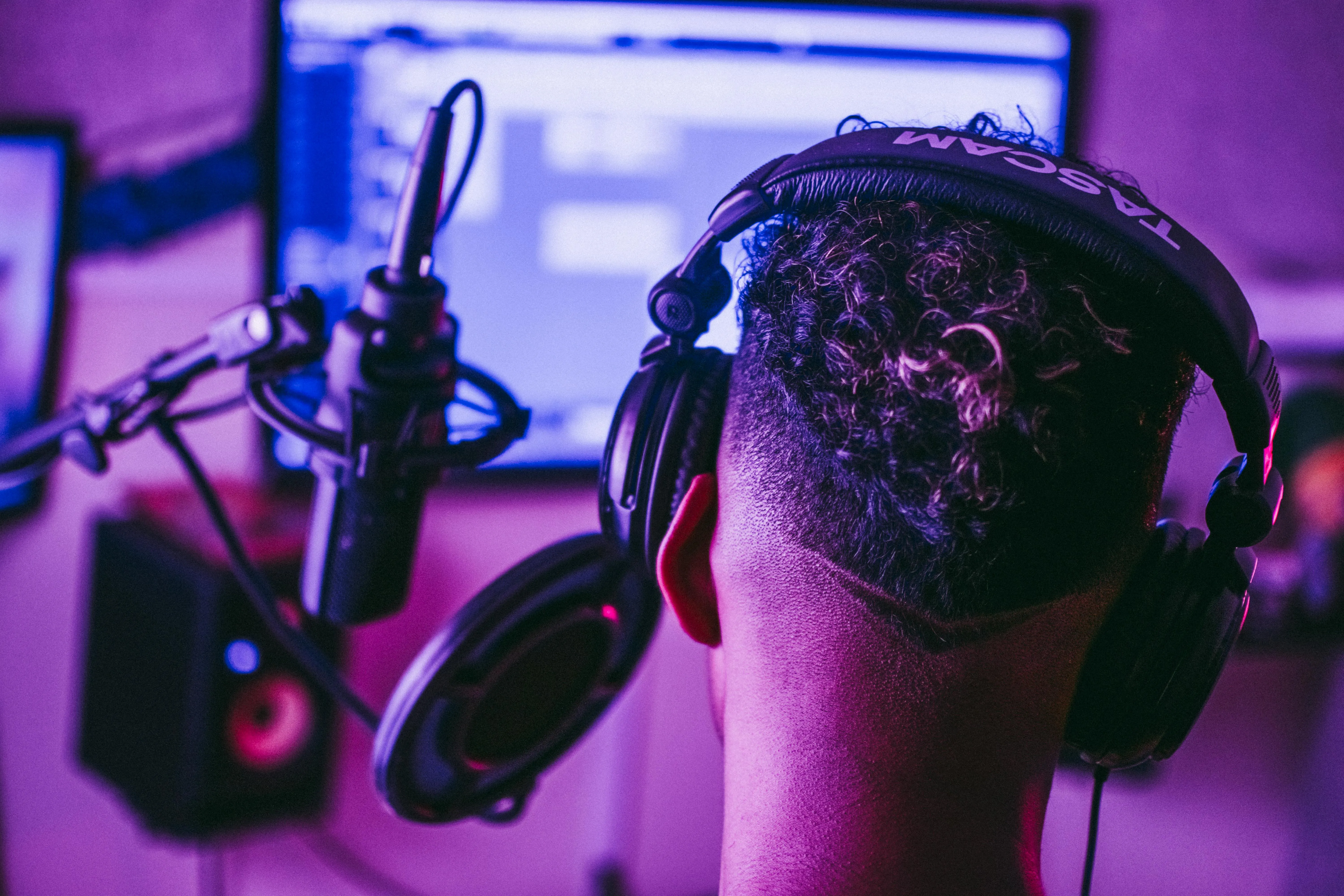 A person with headphones on facing away from the viewer toward a computer, recording vocals into a microphone