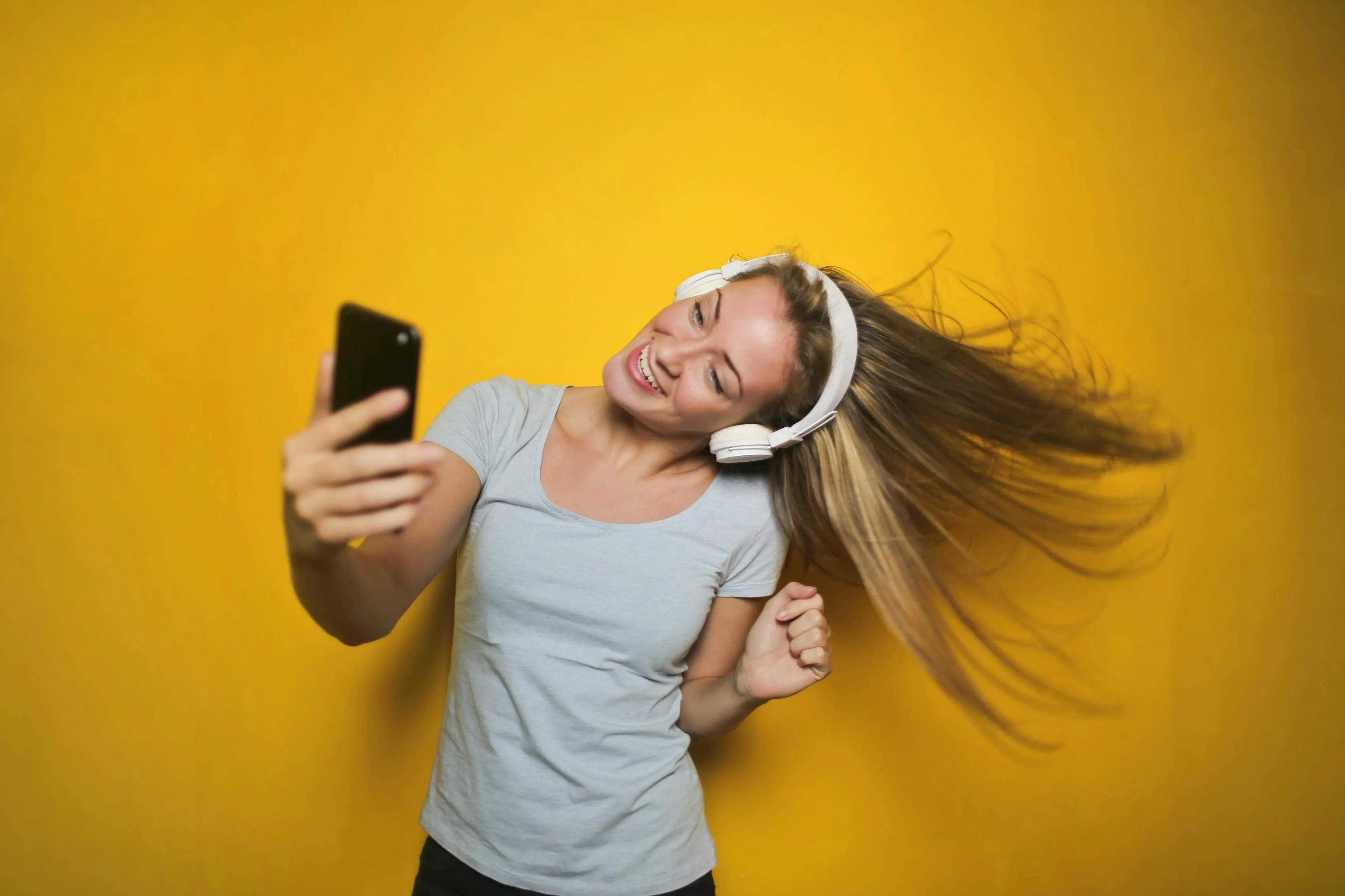 A woman filming herself with a smartphone as she smiles and listens to music on headphones.