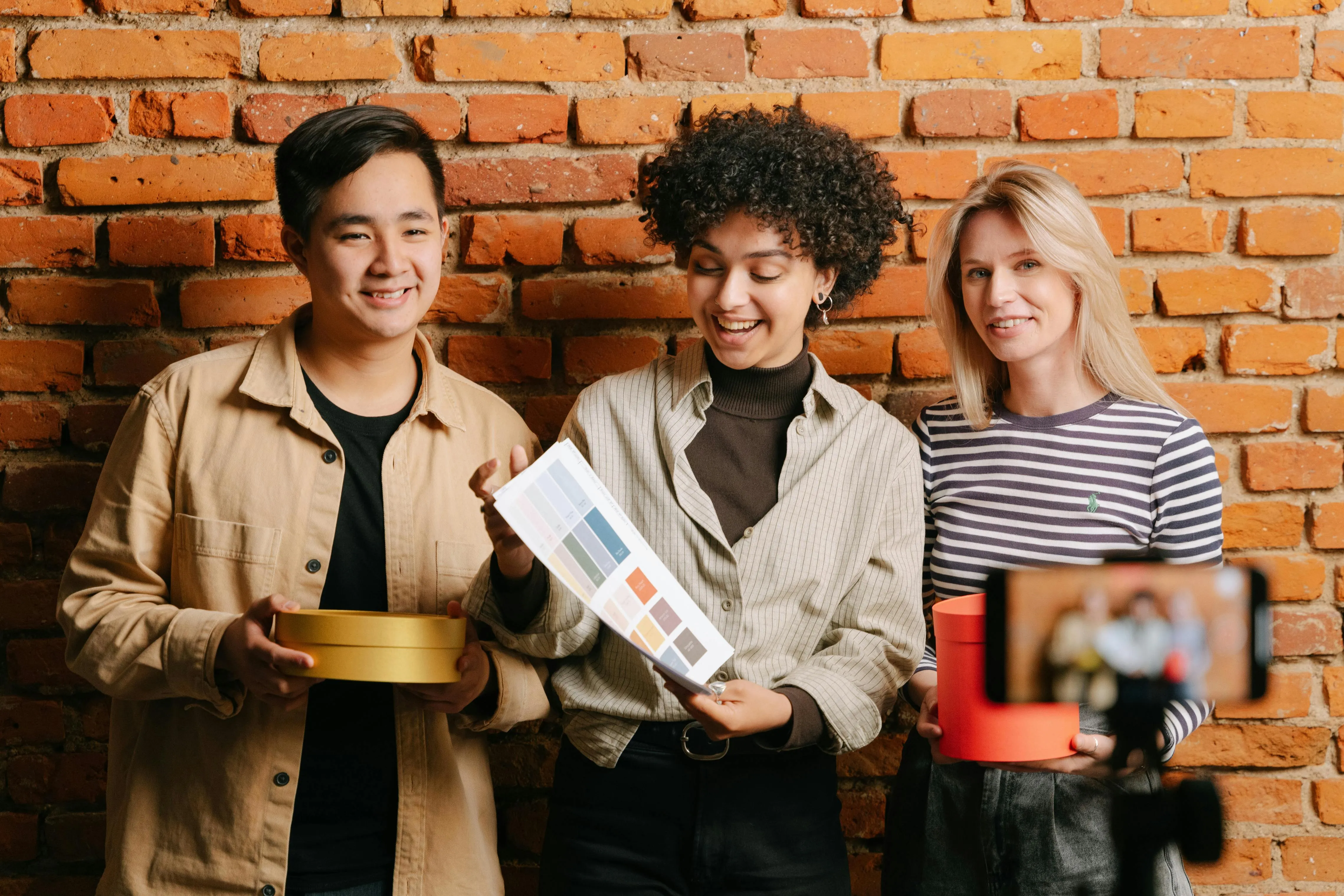 Three members of a marketing team showcasing products to a filming smartphone against a brick background.