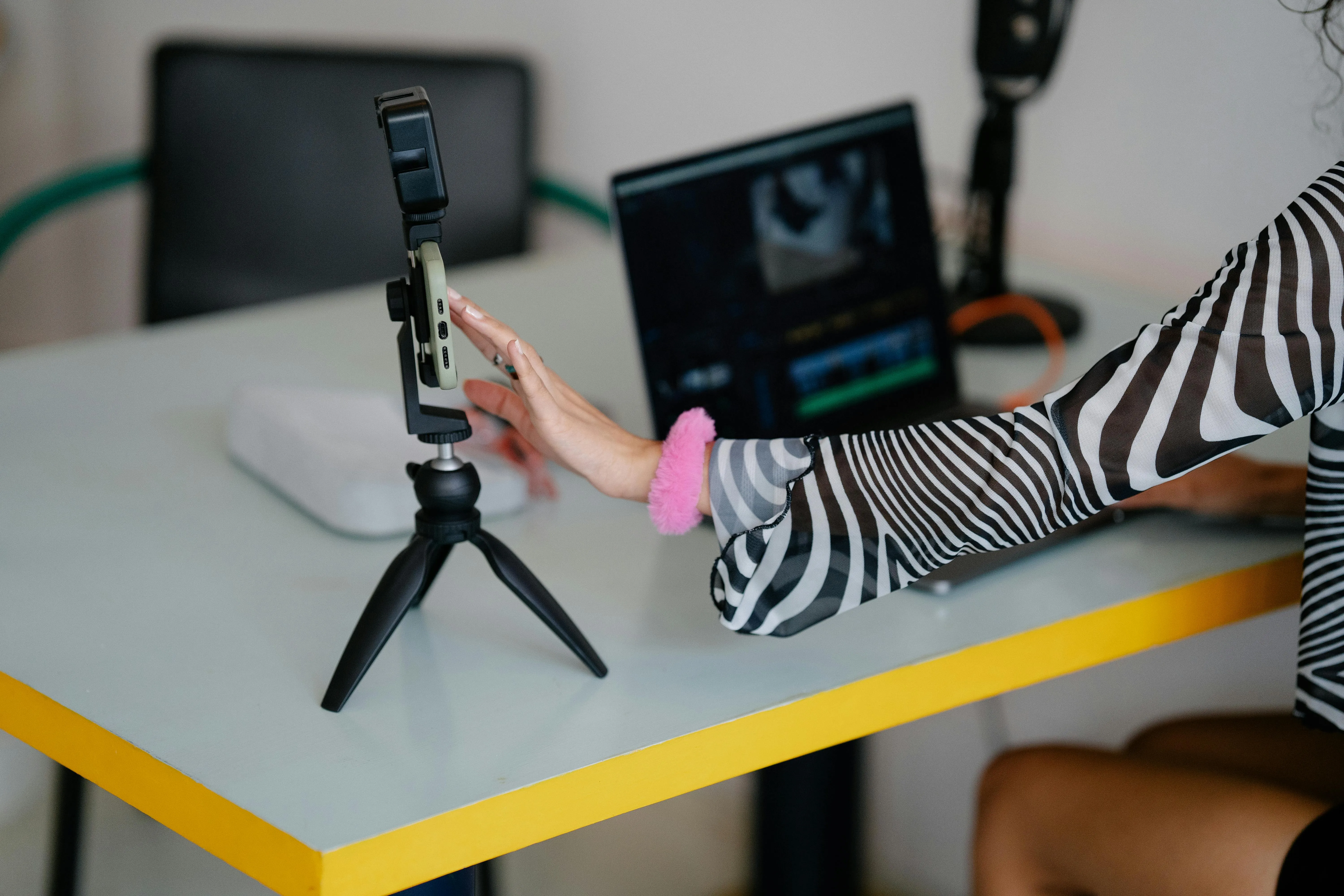 Someone using a smartphone and laptop to record and edit video at a desk.