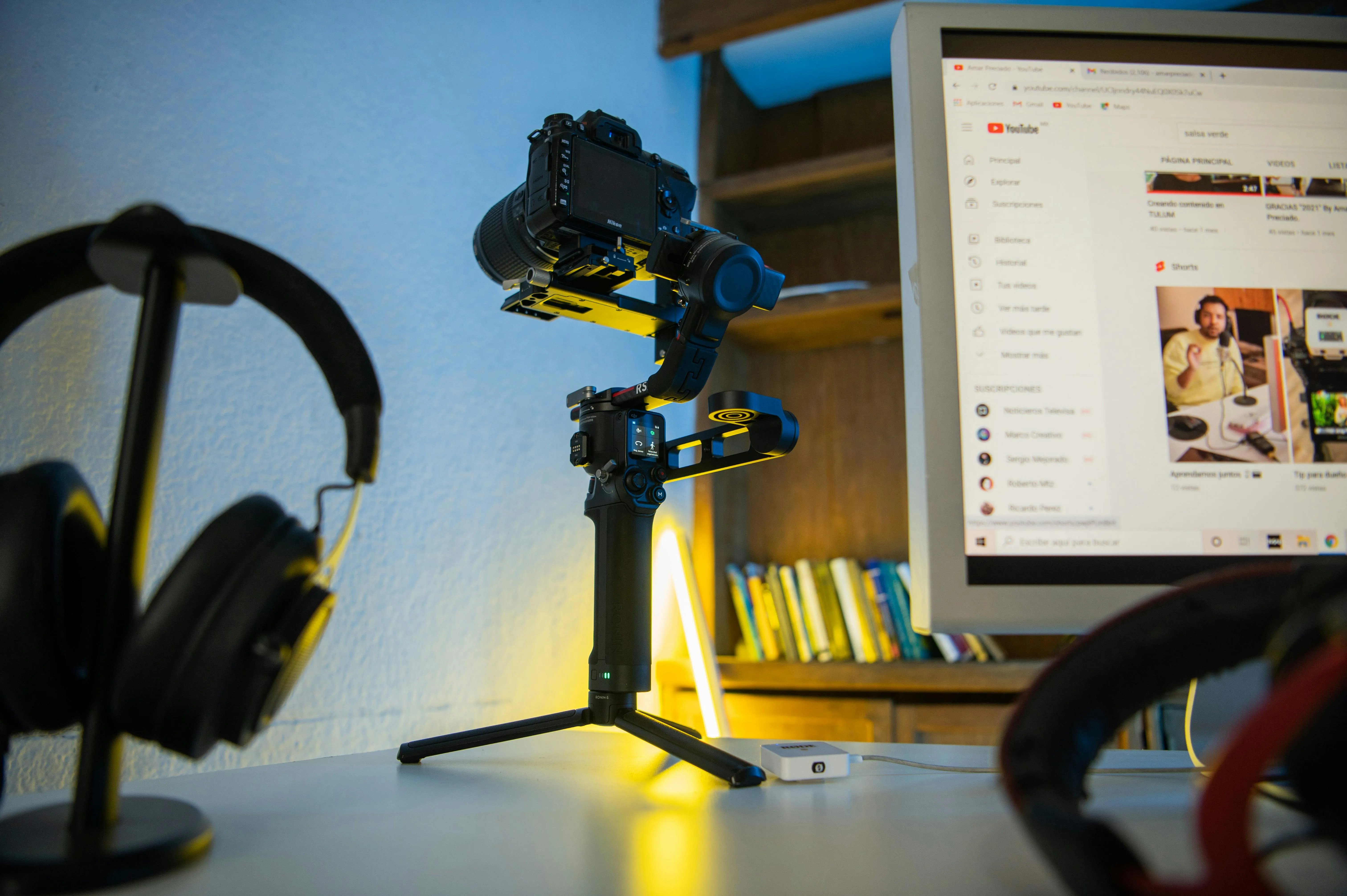 A tripod camera set up on a desk beside headphones and a computer open to YouTube.