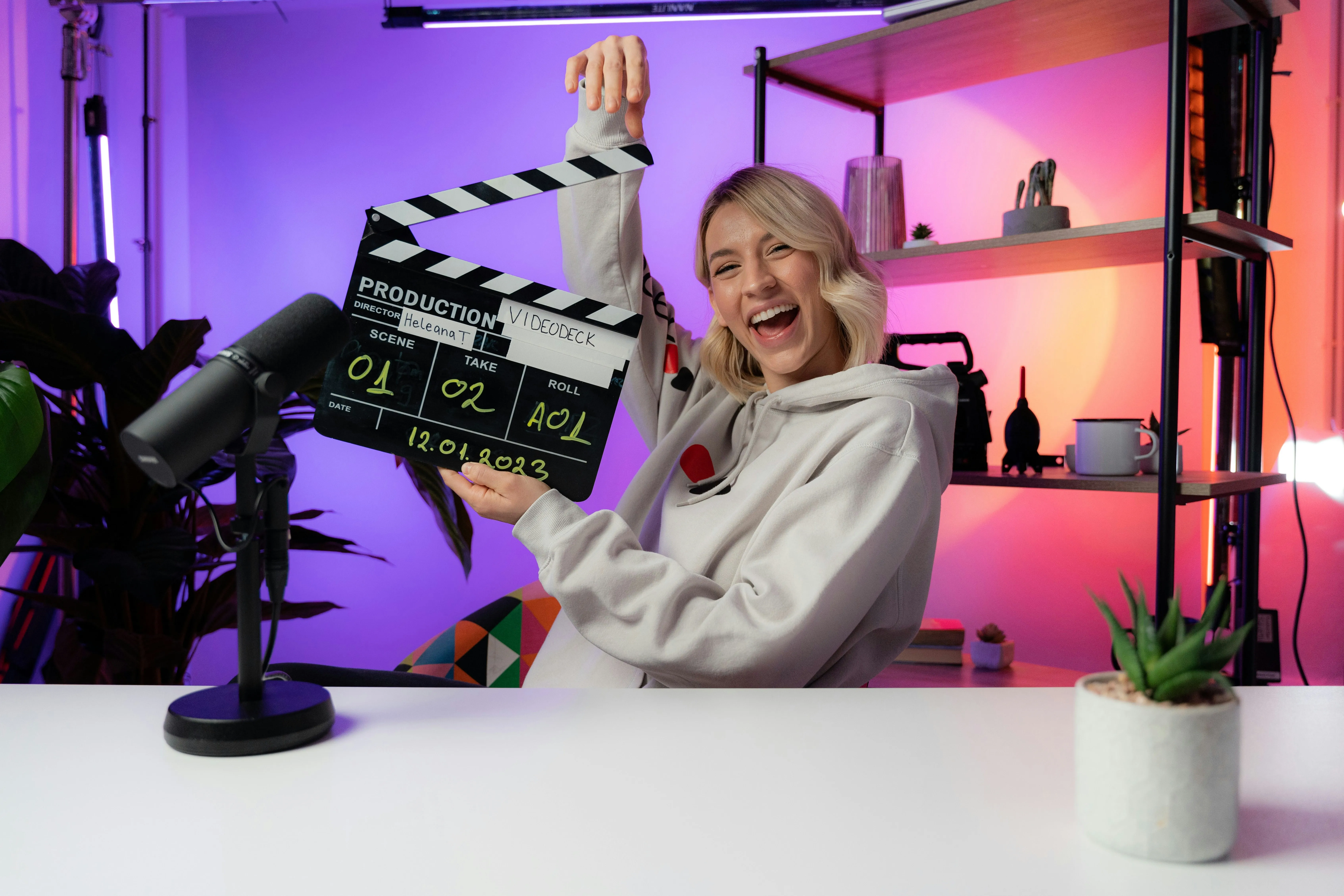 A woman at a desk smiling and holding a clapperboard.